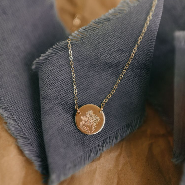 Gold circle floral stamped necklace on a gold chain lays over a blue piece of linen ribbon