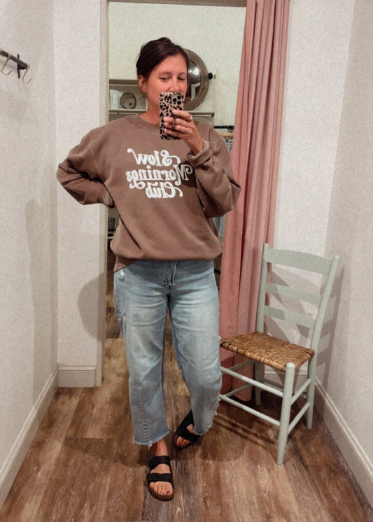 Person taking a mirror selfie wearing a brown sweatshirt with "Slow Mornings Club" and jeans in a room with a pink curtain and white chair.