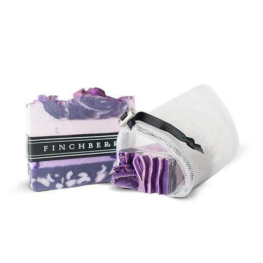 White mesh loofah pouch with black draw string laying on its side with a purple finchberry soap bar sitting next to it and part of the soap bar in the pouch. Against solid white backdrop