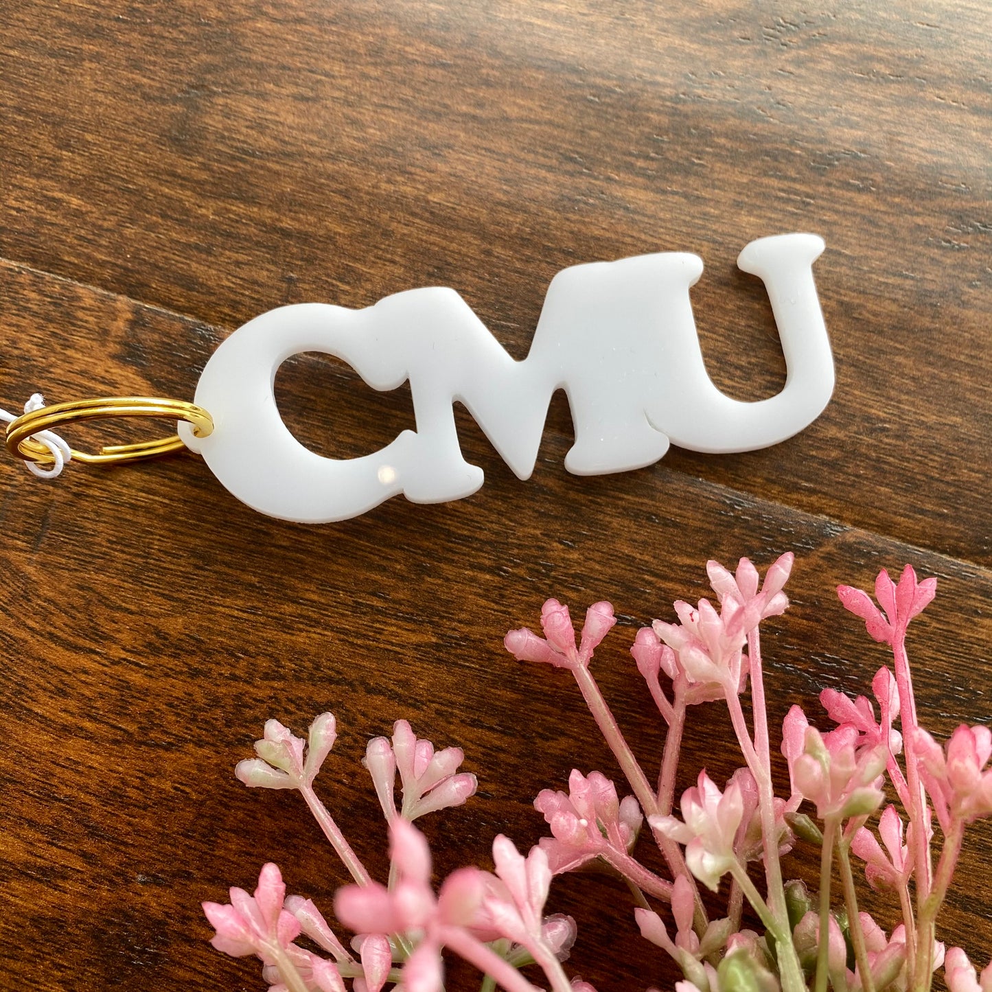 CMU initials in bold font make up white acrylic keychain. It is attached to gold key ring and sitting on a wood surface next to pink florals