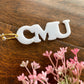 CMU initials in bold font make up white acrylic keychain. It is attached to gold key ring and sitting on a wood surface next to pink florals
