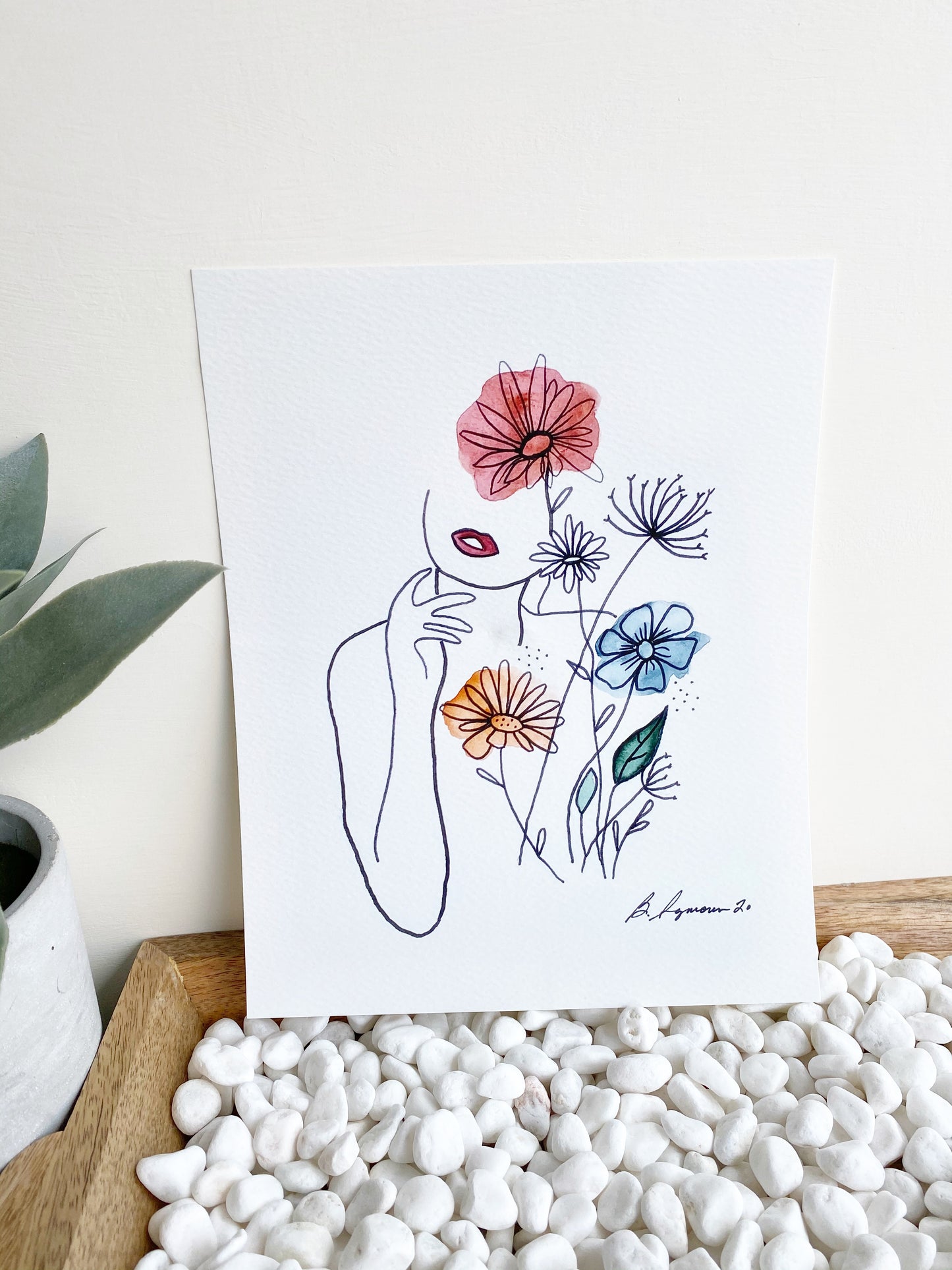 Art print of outline of a woman's face and arm as well as a floral arrangement with watercolor accents. Print sits on wooden tray filled with white pebbles against white wall