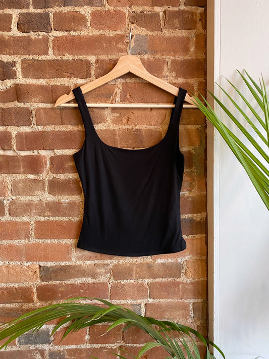 Black square neck tank top hangs on wooden clothes hanger against a brick wall surrounded by greenery